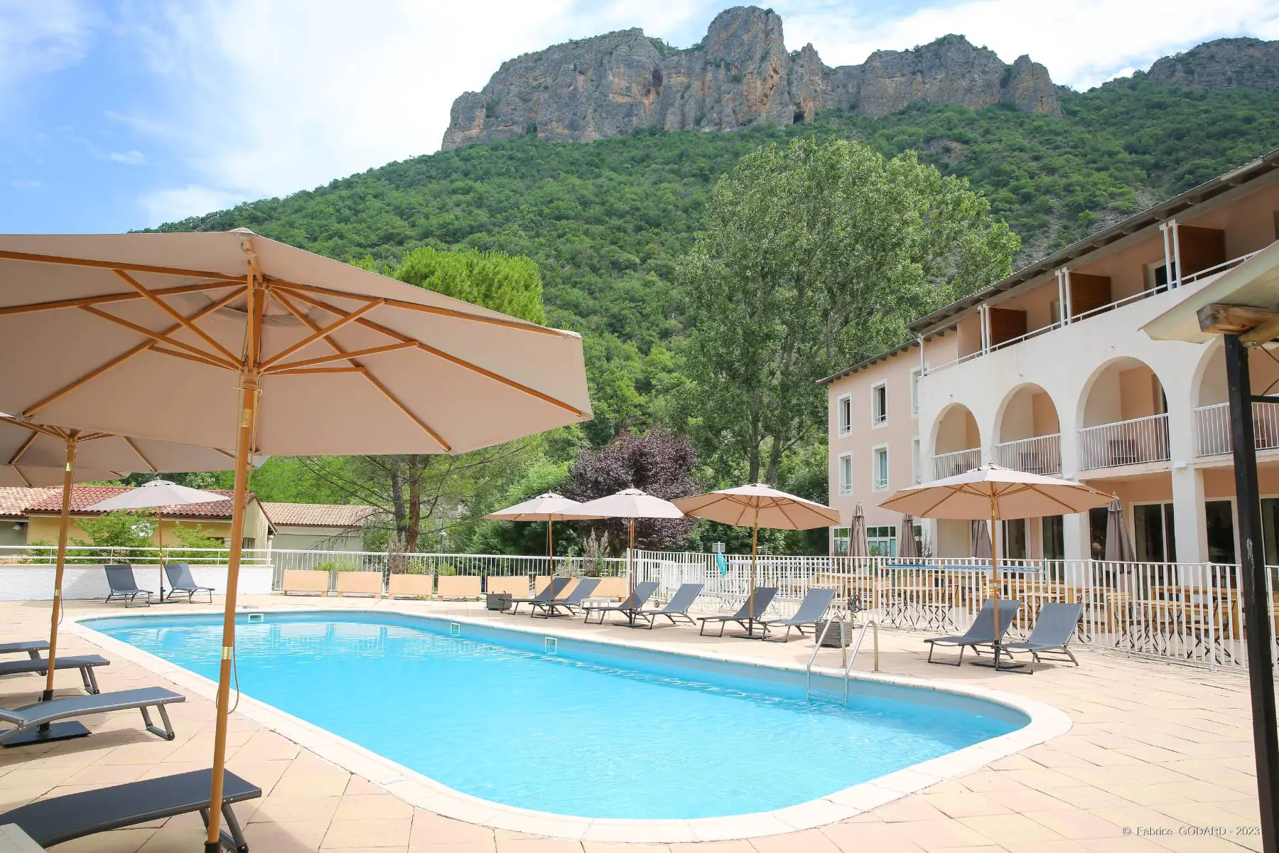 en::Blue swimming pool bordered by a tiled deck, with parasols and sun loungers, offering a view of the mountains. fr::Piscine aux eaux bleues bordée d’une plage carrelée, avec parasols et transats, offrant une vue sur les montagnes.