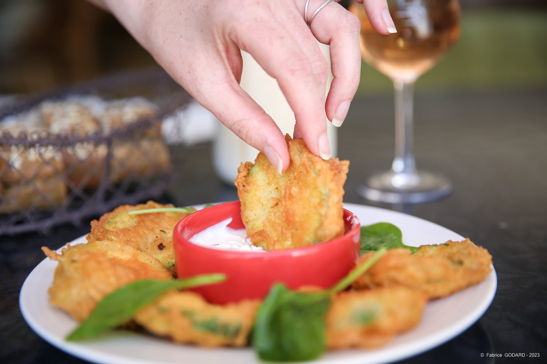 en::Plate of fritters served with herb dipping sauce, glass of wine and bread basket in the background. fr::Plat de beignets accompagnés d’une sauce aux herbes, verre de vin et corbeille de pain en arrière-plan.