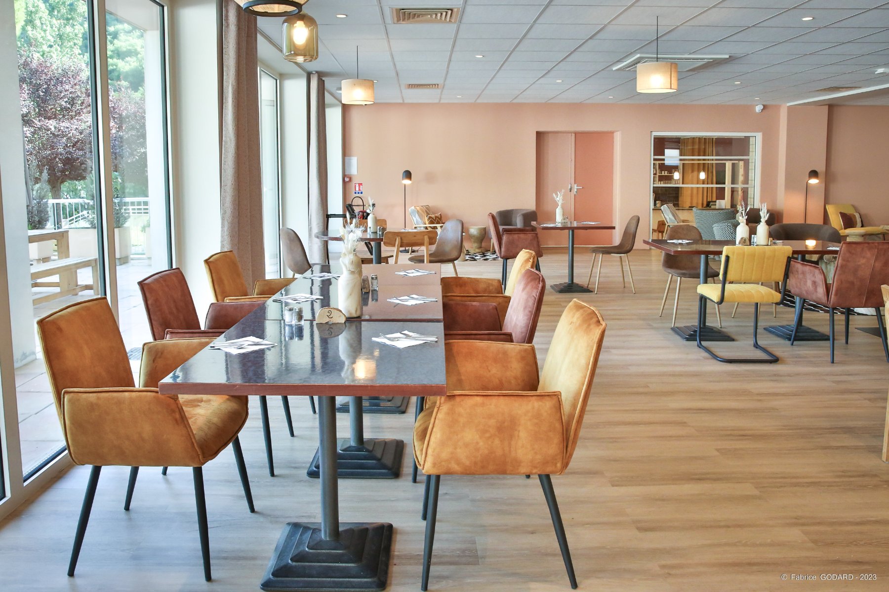 en::Restaurant dining room with large windows opening onto the terrace, orange, yellow and brown chairs, square tables for two, glass wall opening onto the bar. fr::Salle de restaurant avec baies vitrées donnant sur la terrasse, chaises orangées, jaunes et marron, tables carrées pour deux, ouverture vitrée sur le bar.