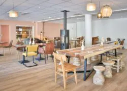 en::Large wooden table set near a stove, surrounded by various tables and chairs, opening onto a cozy and inviting lounge area. fr::Grande table en bois installée près d’un poêle, entourée de tables et de chaises variées, s’ouvrant sur un espace lounge chaleureux et convivial.