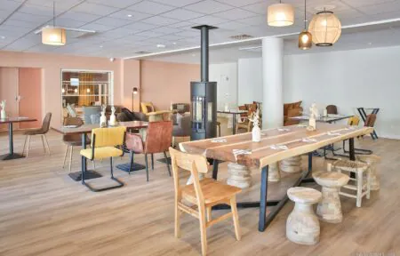 en::Large wooden table set near a stove, surrounded by various tables and chairs, opening onto a cozy and inviting lounge area. fr::Grande table en bois installée près d’un poêle, entourée de tables et de chaises variées, s’ouvrant sur un espace lounge chaleureux et convivial.