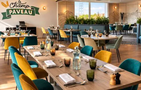 Grande table en bois au premier plan, tables carrées pour 2 personnes le long de banquettes, et tables rondes de 6 à 8 personnes autour.