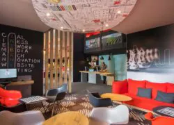 Un lobby d'hôtel avec au premier plan un espace salon avec fauteuils et banquette rouges et noirs, et à l'arrière plan un bureau d'accueil avec deux réceptionnistes.