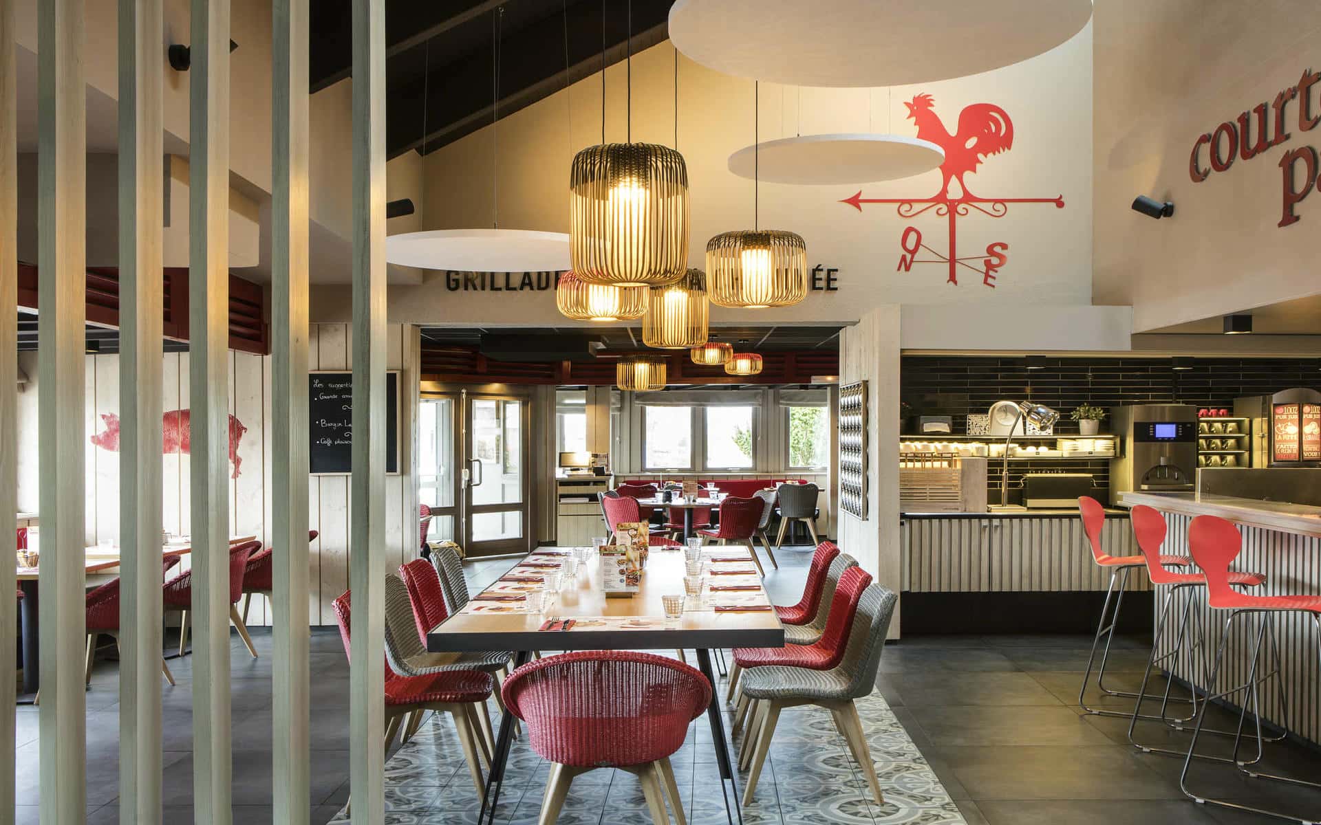 Une salle de restaurant dans les tons rouges, gris et blancs, avec au centre une table dressée dressées pour 10 personnes et à gauche des tabourets alignés le long d'un bar et au fond l'espace buffet du restaurant, surmontée au mur par une grande décoration en forme de girouette avec un coq rouge.