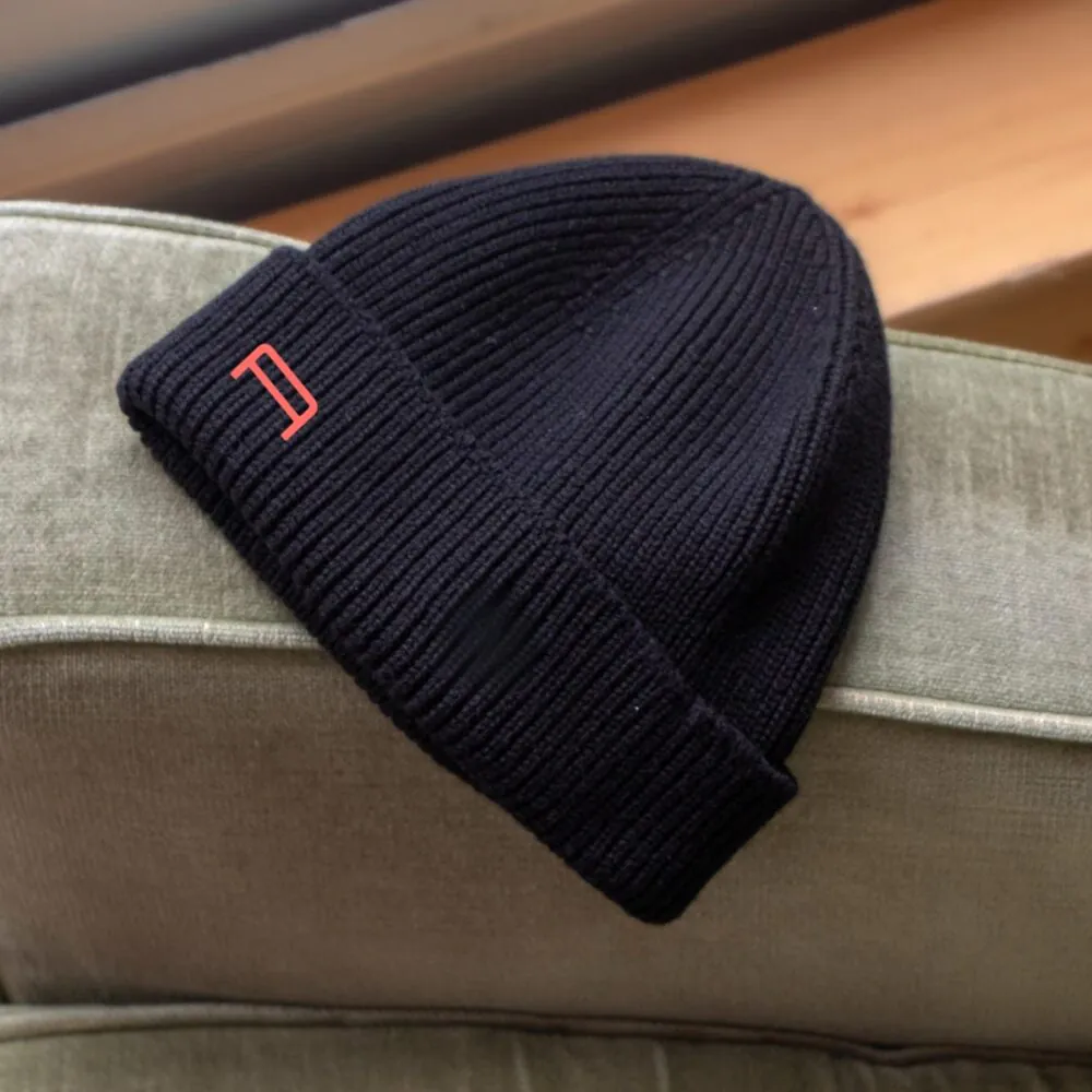 Gros plan d'un bonnet en tricot côtelé bleu marine avec un petit logo orange, objet dérivé hôtel pour séminaire, posé sur un fauteuil en velours vert sauge au premier plan avec un escalier en bois flou à l'arrière-plan.