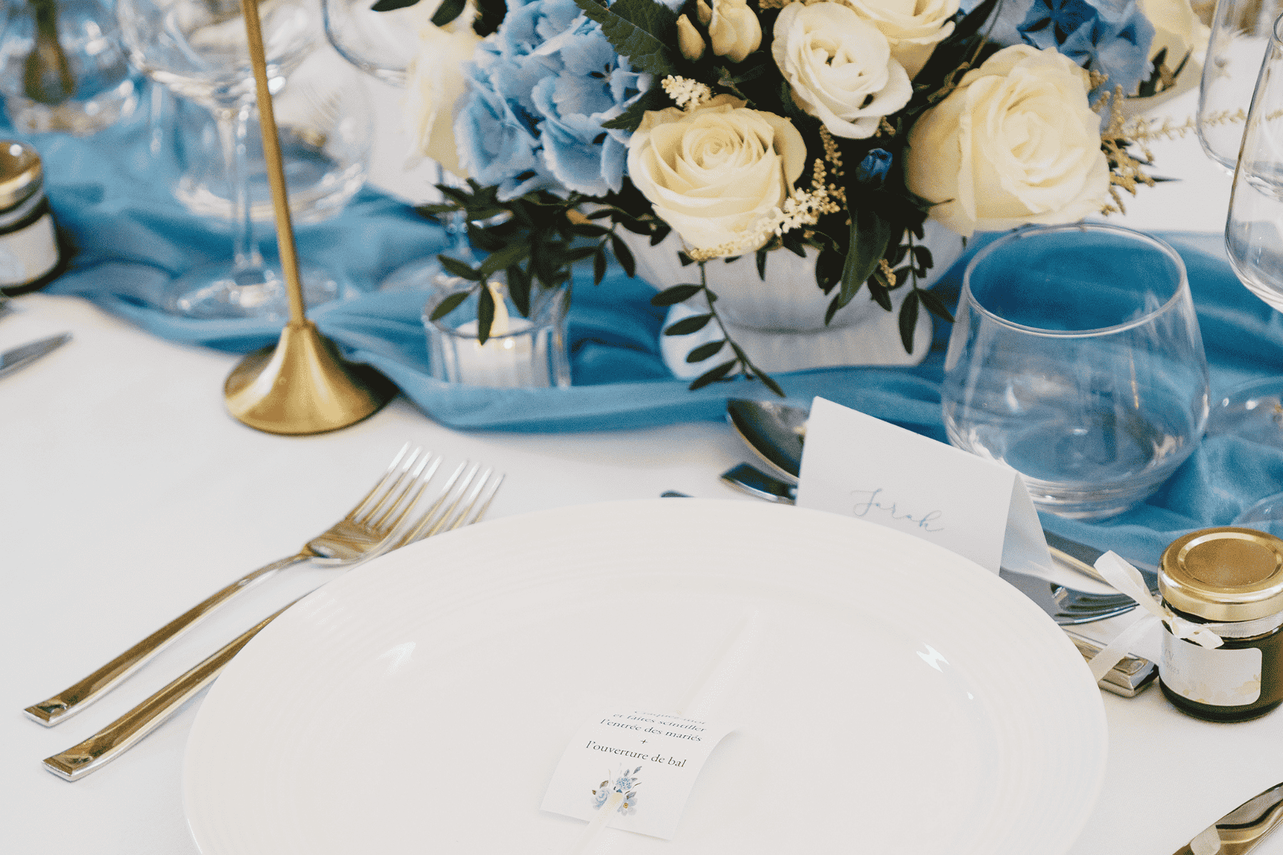 Salle de réception de mariage avec tables nappées de blanc, bouquets bleus, blancs et verts, petits pots de confiture et chemin de table bleu brodé des lettres A et V.