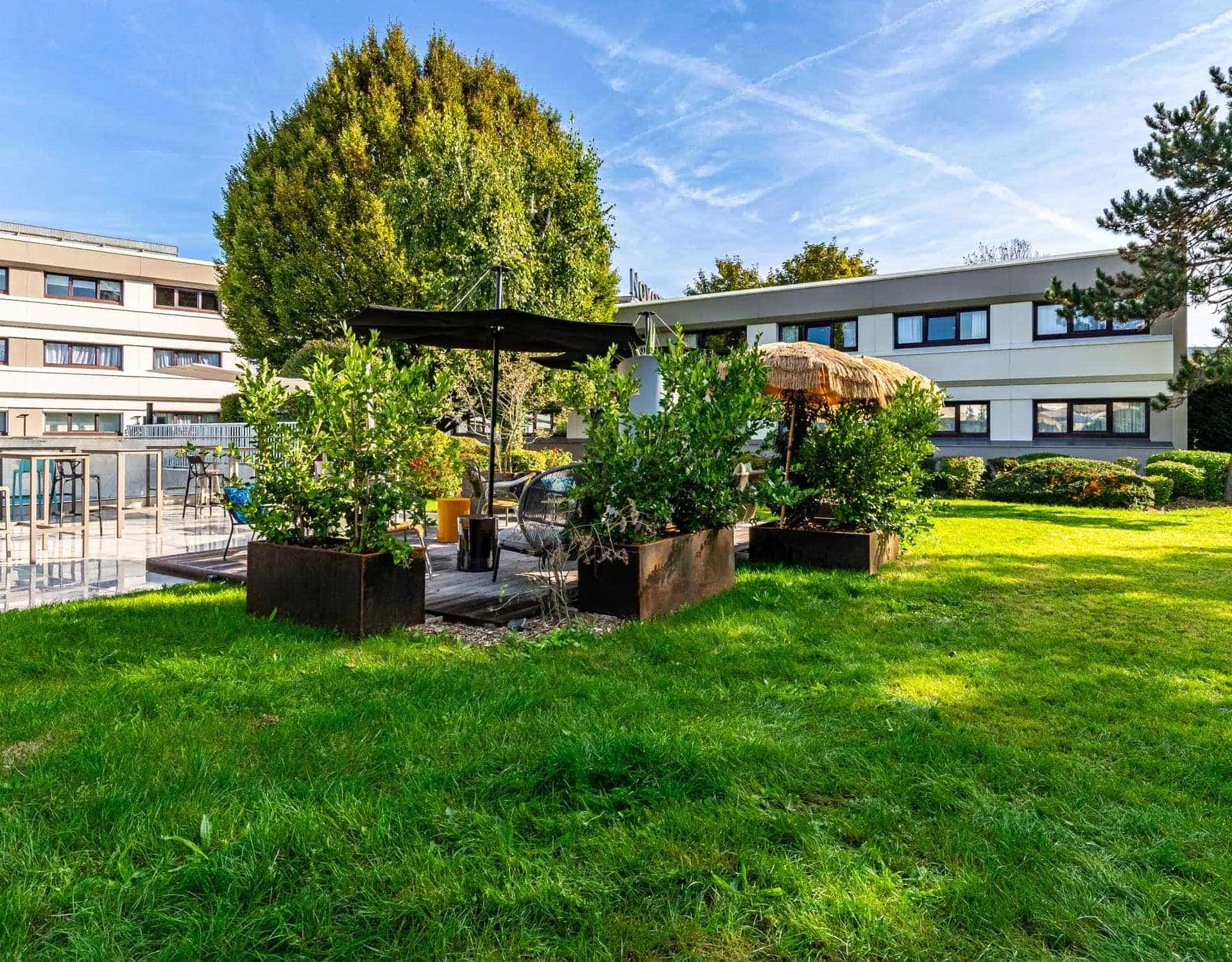 Hotel garden with a large lawn in the foreground, centered around a landscaped wooden deck furnished with outdoor seating and shaded by canopies; low-rise hotel buildings and mature trees frame the background.