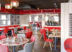 Une salle de restaurant dans les tons rouges, gris et blancs, avec de nombreuses tables dressées pour 2 à 6 convives, avec une séparation ouverte au milieu où sont alignées en décoration 7 bouteilles de champagne de taille décroissante.