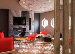 Un espace salon moquetté avec 4 tables basses entourées de fauteuils et banquette dans les ton rouges, gris et noir, avec au fond une télébision au mur.