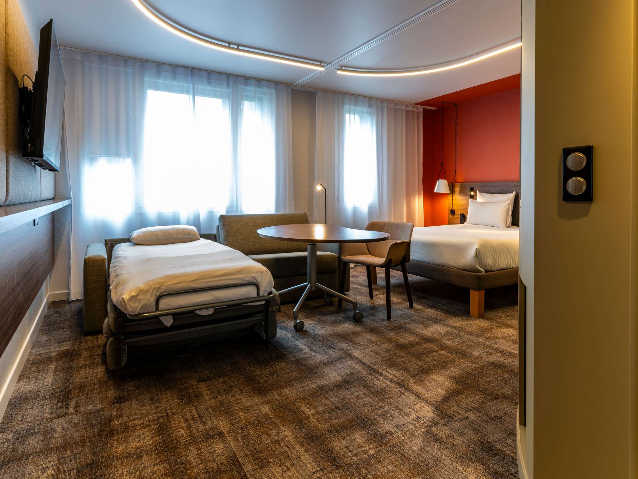 Suite with brown checkered carpet, red walls, a double bed and a partially opened sofa bed: one bed made up and one sofa area preserved. The ceiling features a rounded LED light encircling the sleeping area.