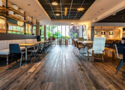 Une très grande salle de petit déjeuner avec, de part et d’autre d’une allée centrale, des banquettes et tables ou des tables et chaises. En ligne de mire, de larges baies vitrées donnant sur un espace extérieur végétalisé. Un espace lumineux, volumineux et contemporain, avec une hauteur sous plafond très agréable.