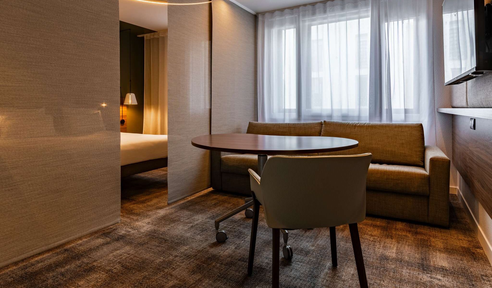 Suite with brown checkered carpet, brown walls, a large double bed and a two-person sofa bed in front of a stylish rolling wooden table, separated from the sleeping area by sleek brown blinds.