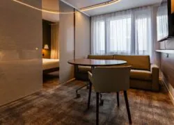Suite avec moquette à carreaux marron, murs marron, large lit double et canapé-lit deux personnes devant une table roulante design en bois, séparée du coin nuit par des stores marron épurés.