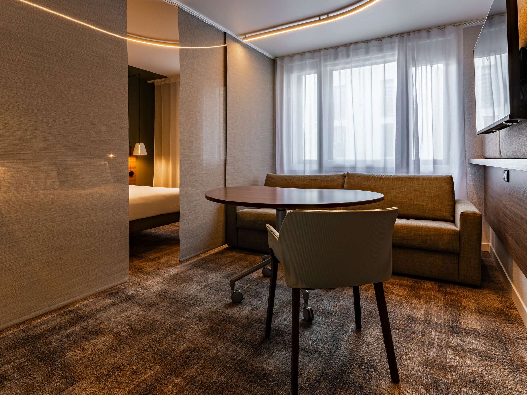 Suite with brown checkered carpet, brown walls, a large double bed and a two-person sofa bed in front of a stylish rolling wooden table, separated from the sleeping area by sleek brown blinds.