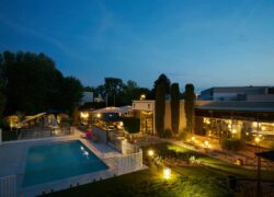 A park illuminated at night featuring, from left to right, the swimming pool, the restaurant terrace, and the hotel’s low-rise building.