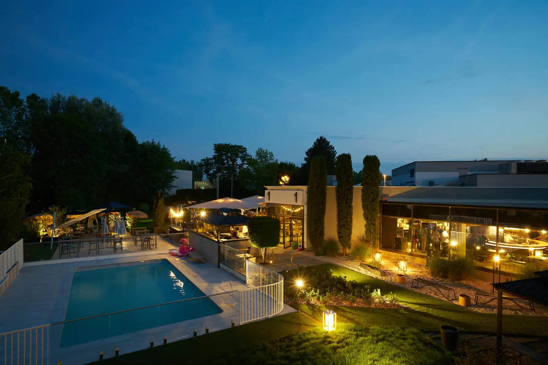 A park illuminated at night featuring, from left to right, the swimming pool, the restaurant terrace, and the hotel’s low-rise building.
