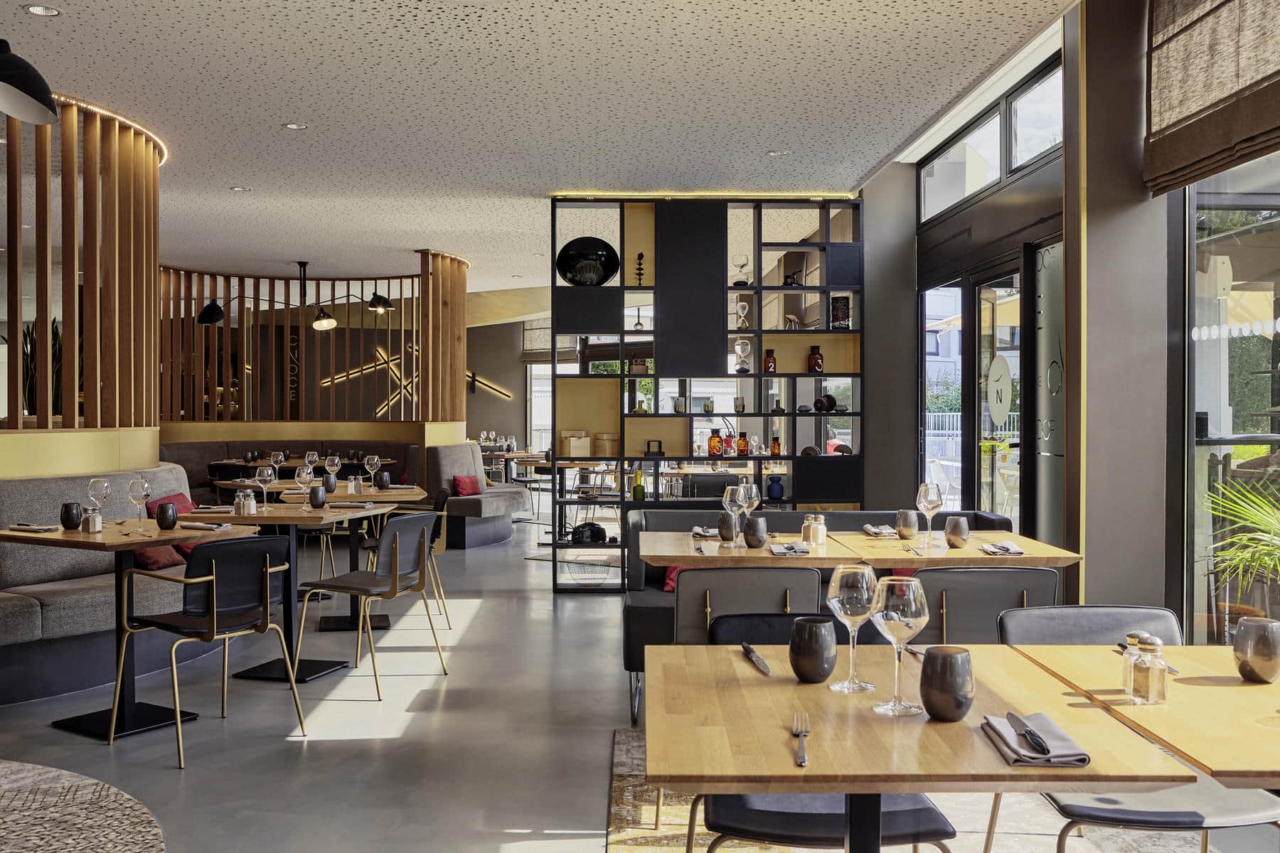 A modern and welcoming restaurant dining room featuring several fully set tables, grey armchairs and banquettes, a large bookshelf-style unit in the centre displaying decorative objects, and an entire right-hand wall made of large bay windows.