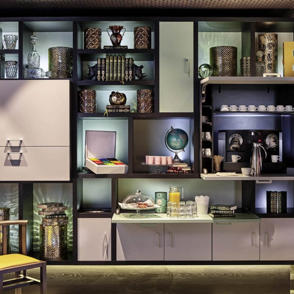 Close-up of a wall section featuring a large bookshelf-style unit in brown and blush pink tones, displaying decorative objects and housing a coffee, tea, and fruit juice station.