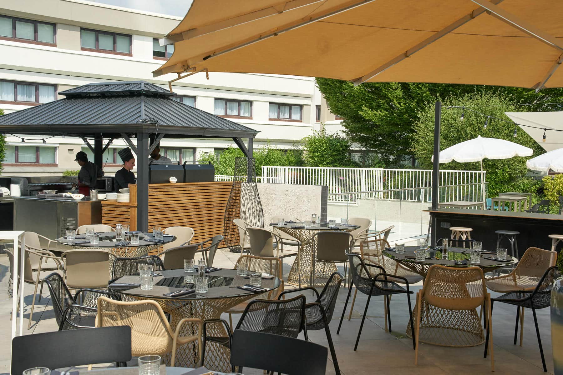 A tiled restaurant terrace with round tables set for six guests, cream-coloured parasols, and a black gazebo in the middle ground where chefs are preparing a barbecue, with a white building and trees visible in the background.