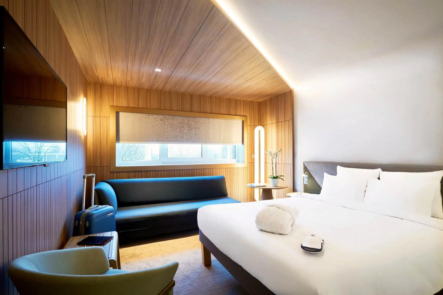 A bright bedroom with a large double bed on the right facing a wall-mounted flat-screen TV, and at the back of the room a black sofa beneath a large window.