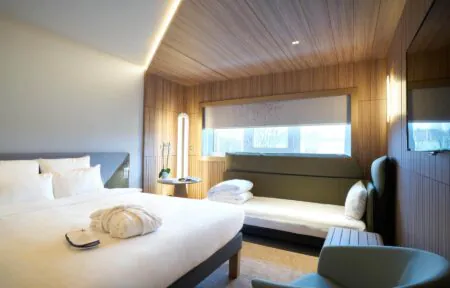 A bright hotel room with a large double bed on the left facing a wall-mounted television screen, and at the back a sofa bed beneath a large window.