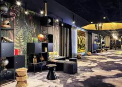 Focus sur un côté d'un vaste hall d'accueil très coloré à la décoration contemporaine avec une moquette noir et blanche, un plafond noir, du mobilier moderne noir et marbre clair, des éléments de décoration dorés et quelques pans de murs rayés noir et blancs et d'autre présentant un décor de forêt tropicale.