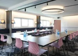 Une salle de réunion lumineuse dressée en U pour 16 personnes avec du mobilier contemporain, des chaises texturées roses, noires et grises et une moquette noir et blanches.