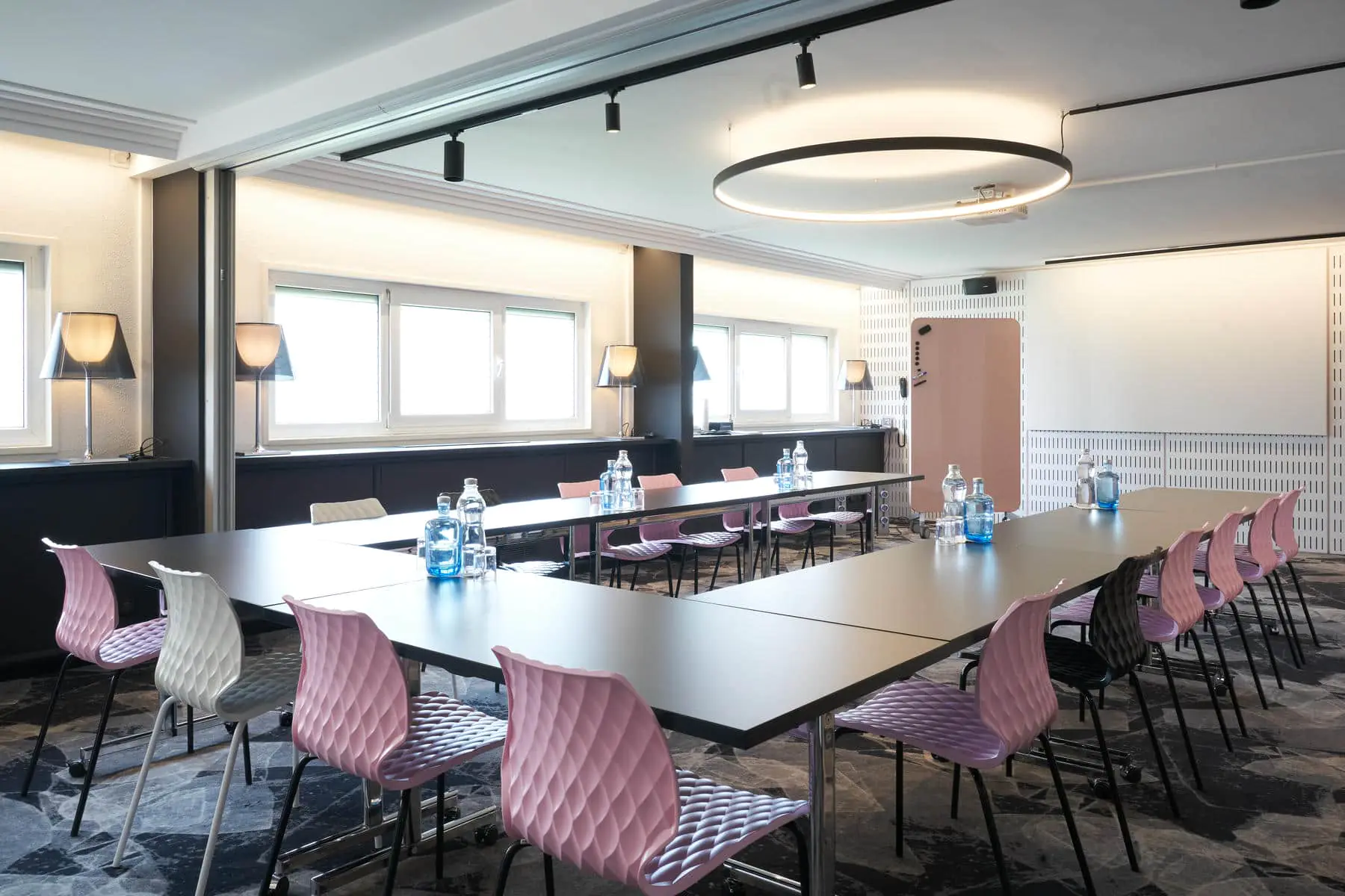 A bright meeting room set up in a U-shape for 16 people, featuring contemporary furniture, textured pink, black, and grey chairs, and a black-and-white carpet.