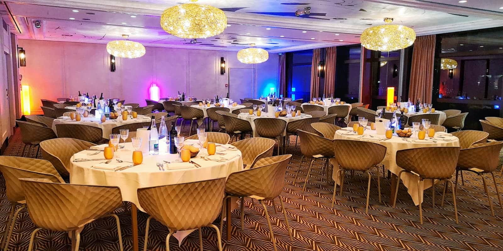 A reception room at night featuring round tables dressed and set for a group gala dinner, with festive lighting throughout the space.