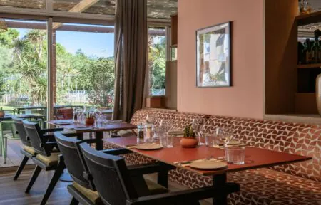 Une salle de restaurant dans les tons roses et verts avec quelques tables dressées donnant sur une baie vitrée avec vue sur terrasse et parc.
