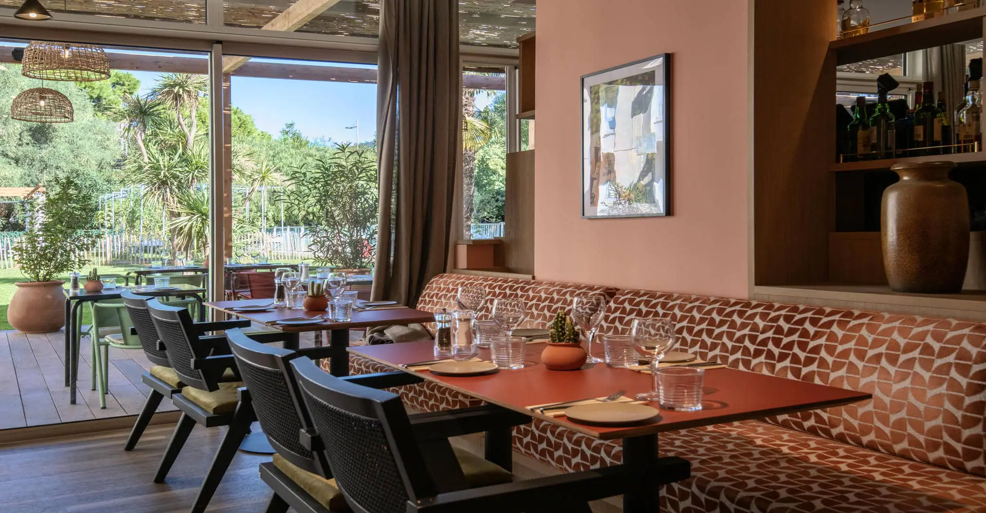 Une salle de restaurant dans les tons roses et verts avec quelques tables dressées donnant sur une baie vitrée avec vue sur terrasse et parc.