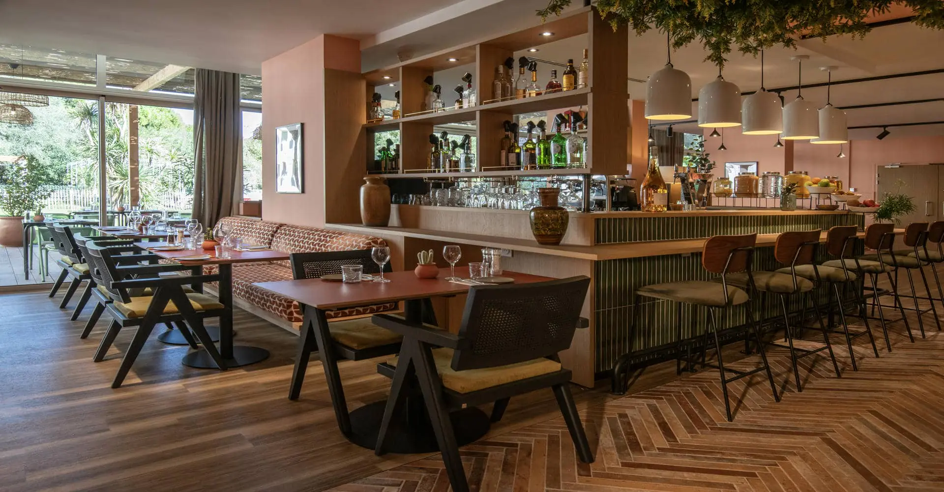 Une salle de restaurant dans les tons roses et verts avec quelques tables dressées sur la gauche donnant sur une baie vitrée avec vue sur parc et un bar central avec des tabourets de bar alignés sur la droite.
