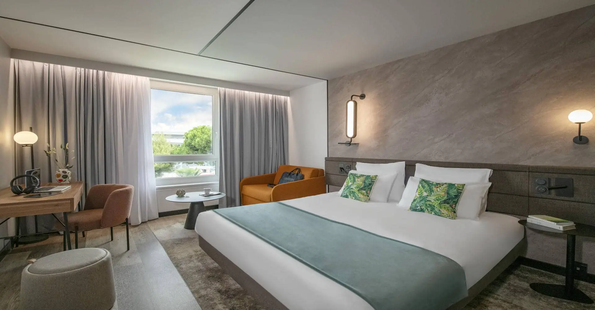 Une chambre d'hôtel spacieuse avec grand lit double au premier plan, un coin bureau au fond à gauche et une fenêtre donnant sur un parc en arrière plan.