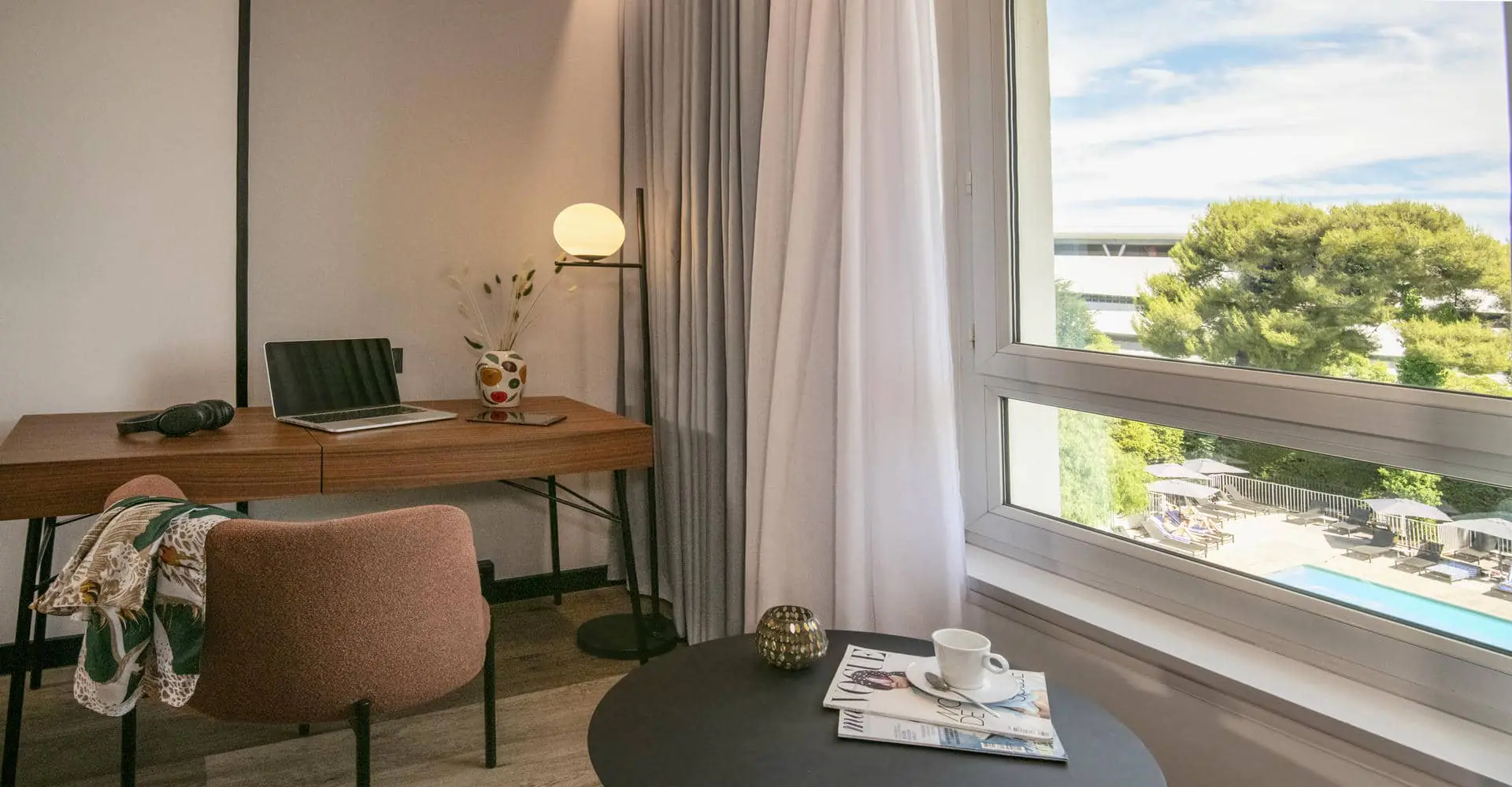 Un coin bureau dans une chambre d'hôtel avec vue par la fenêtre sur une piscine et un parc arboré.