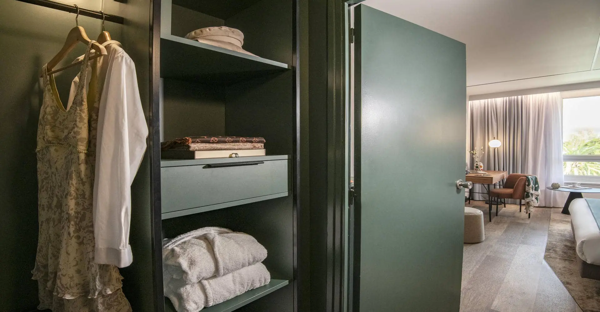 Vue sur une chambre d'hôtel spacieuse avec un dressing ouvert où sont pendus des habits au premier plan et la chambre que l'on devine derrière la porte ouverte en arrière plan.