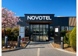Façade d’un hôtel encadrée de plates-bandes arborées et fleuries, avec le logo Novotel au-dessus de la porte d’entrée.