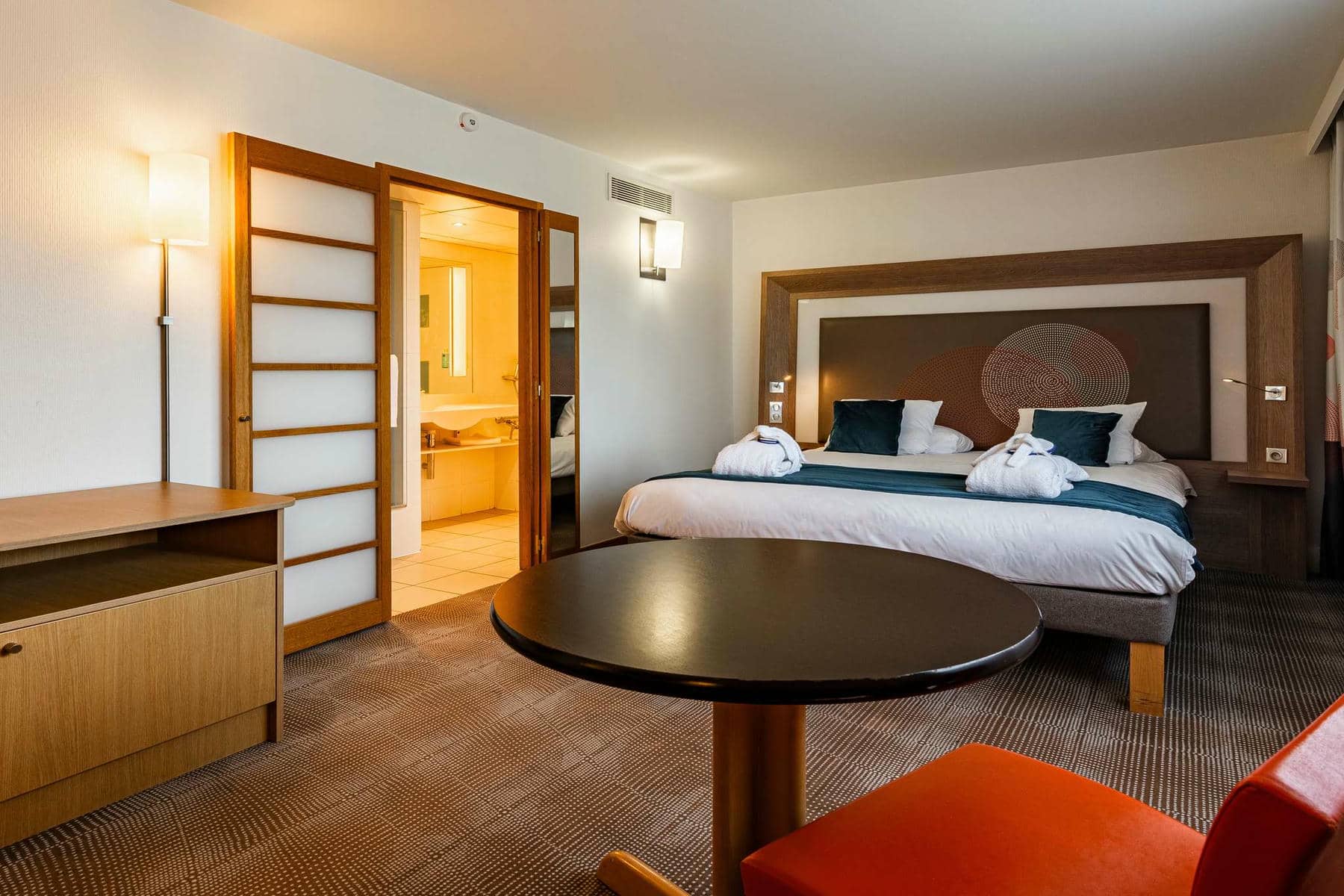 Une grande chambre moquetée avec au premier plan un coin bureau, au second plan une large tête de lit boisée encadrant un grand lit double où sont posés des peignoirs, et qui est attenant à une porte en bois coulissante donnant sur une grande salle de bain.