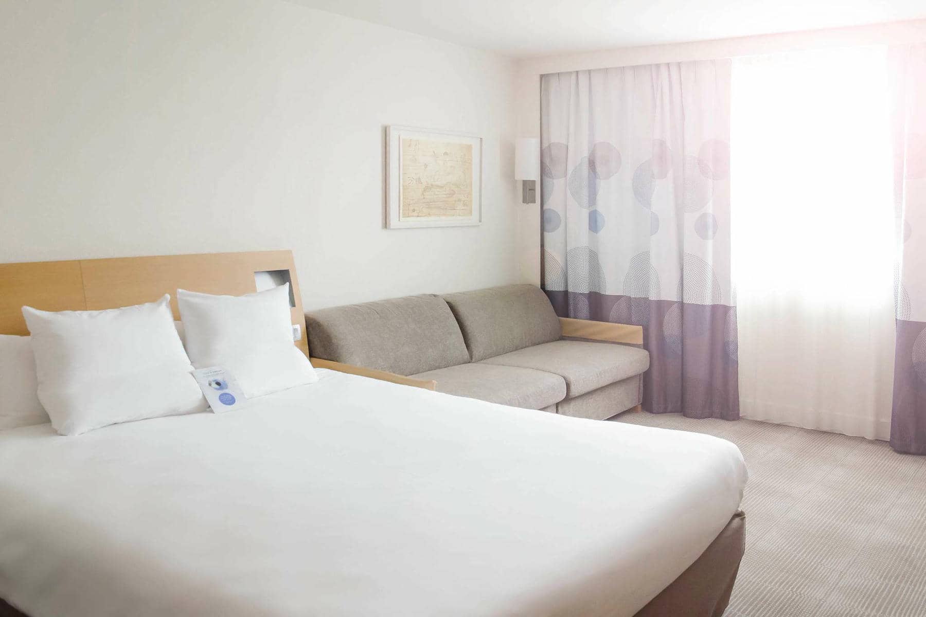 Une chambre lumineuse avec à gauche au premier plan un lit double, une banquette grise surmontée d'un cadre décoratif en second plan, et une fenêtre avec voilage blanc et gris à l'arrière plan.