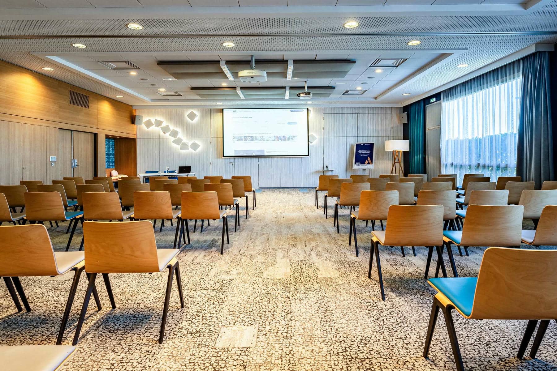 Une salle de réunion moquettée et lumineuse dressée en théâtre avec plusieurs rangées de chaises en bois clair et pieds noirs tournées vers un écran de projection mural, et une large baie vitrée sur la droite.