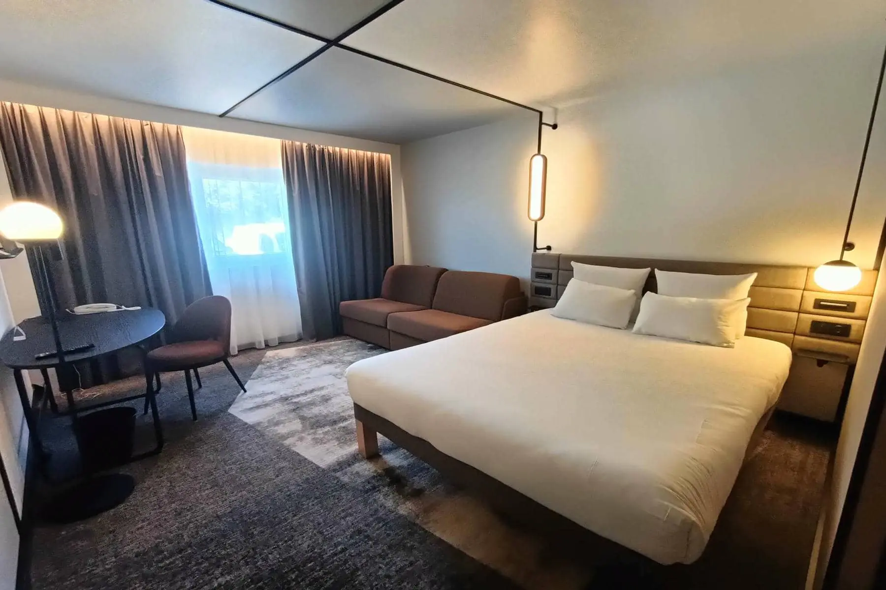 Une chambre avec à droite un lit double, une tête de lit grise molletonnée sur mur blanc, et au second plan un coin bureau sur la gauche et une banquette marron sur la droite, donnant sur une fenêtre avec voilages en arrière plan.