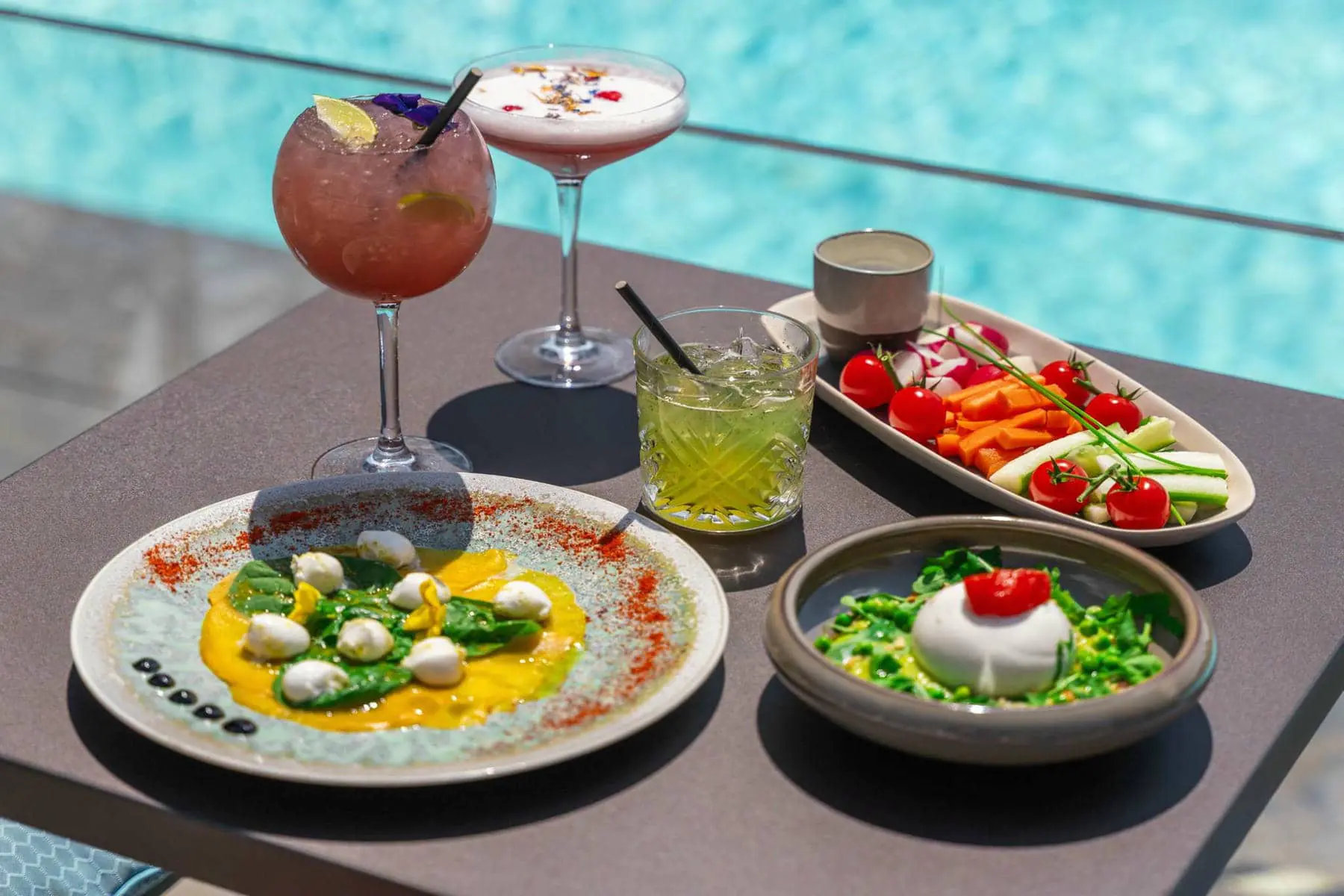 Gros plan sur une petite table carrée grise au bord de la piscine, sur laquelle se trouvent de gauche à droite 3 verres de cocktails différents, une assiette plates de billes de mozzarella et de salade, un plat rectangulaire présentant des crudités variées et une assiette creuse avec une boule de buratta sur lit de légumes verts.