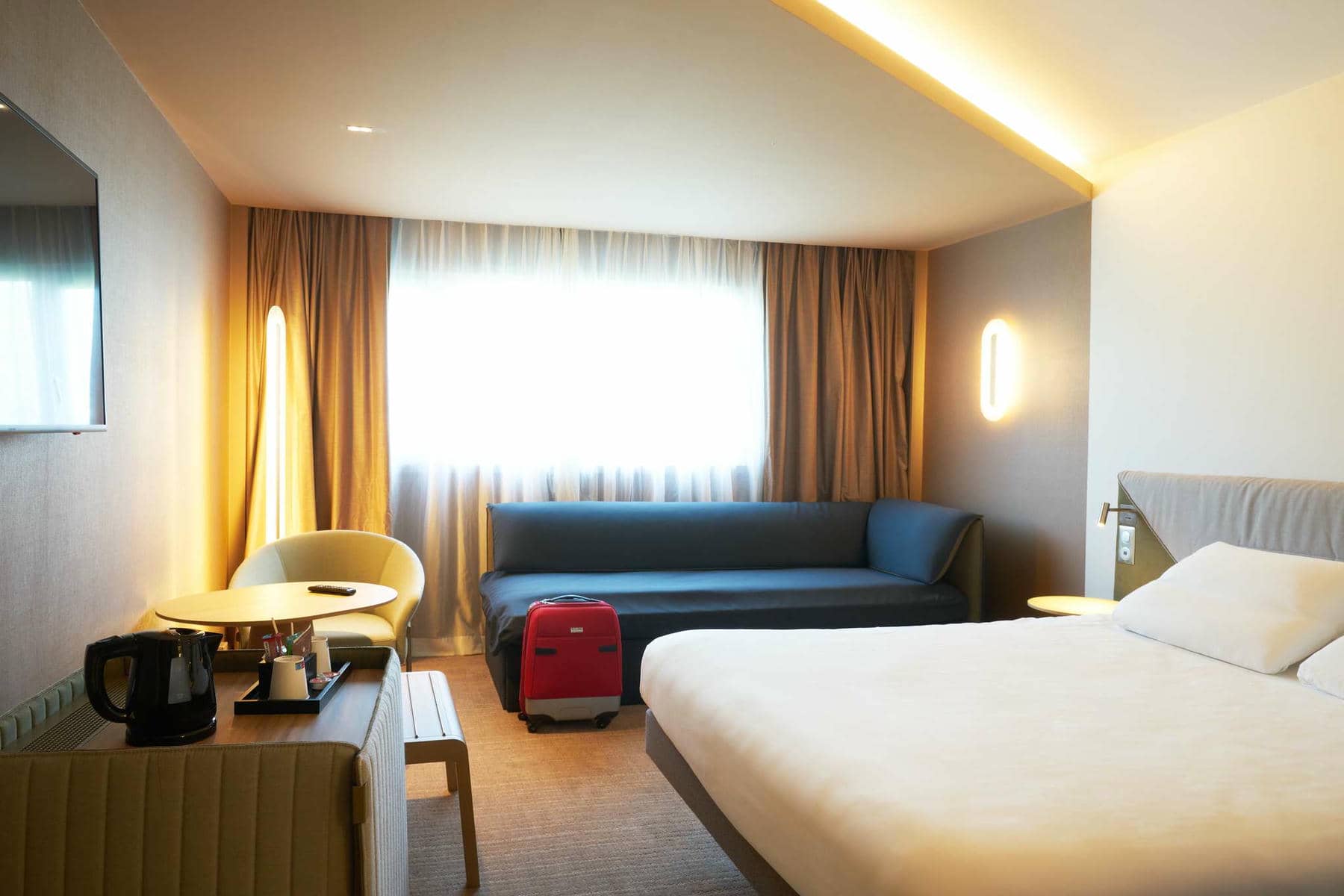 A bright bedroom featuring, in the right foreground, a double bed facing a wall-mounted flat-screen TV, and in the background a workspace on the left and a black banquette on the right beneath a large window with sheer curtains.