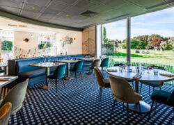 Une salle de restaurant lumineuse aux sièges colorés avec moquette à damiers noir et blanc, et en fond une baie vitrée donnant sur de larges espaces verts.