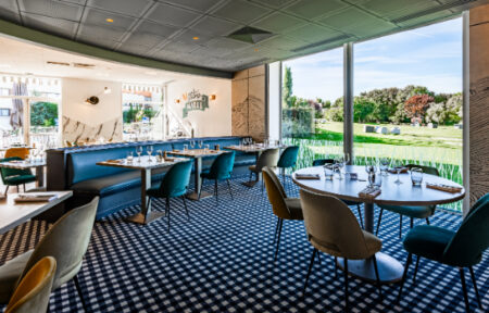 Une salle de restaurant lumineuse aux sièges colorés avec moquette à damiers noir et blanc, et en fond une baie vitrée donnant sur de larges espaces verts