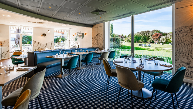 Une salle de restaurant lumineuse aux sièges colorés avec moquette à damiers noir et blanc, et en fond une baie vitrée donnant sur de larges espaces verts.