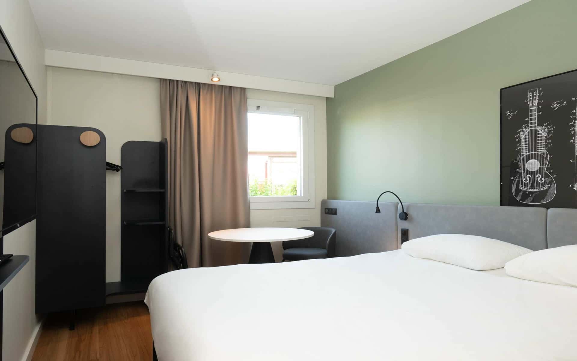 Une chambre d'hôtel avec sur la droite et au premier plan un lit double faisant face à un écran de télévision au mur, au fond de la pièce un petit espace bureau avec une table ronde et un fauteuil et en arrière plan une fenêtre avec vue sur du feuillage.