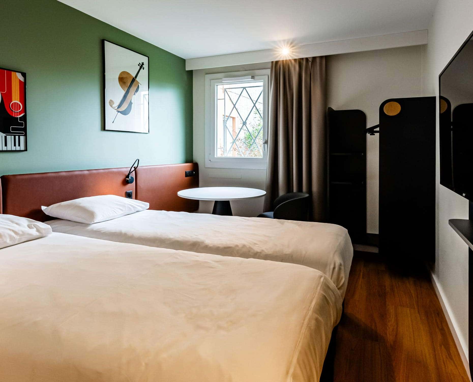 Une chambre d'hôtel avec sur la gauche et au premier plan deux lits simple faisant face à un écran de télévision au mur, au fond de la pièce un petit espace bureau avec une table ronde et un fauteuil et en arrière plan une fenêtre avec vue sur du feuillage.