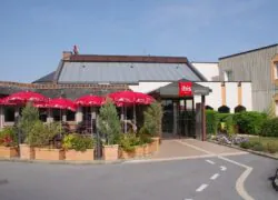 Une façade d'hôtel de deux étages sur un parking avec sur la gauche une terrasse de restaurant avec parasols rouges.