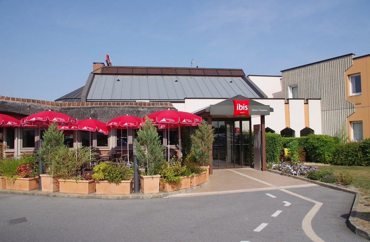 Une façade d'hôtel de deux étages sur un parking avec sur la gauche une terrasse de restaurant avec parasols rouges.