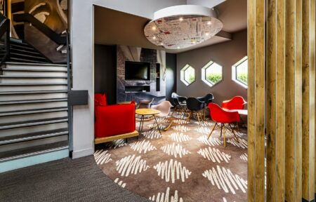 Un coin salon avec des chaises, des tables basses et une banquette rouge, délimité par une moquette marron à damier rayé blanc.